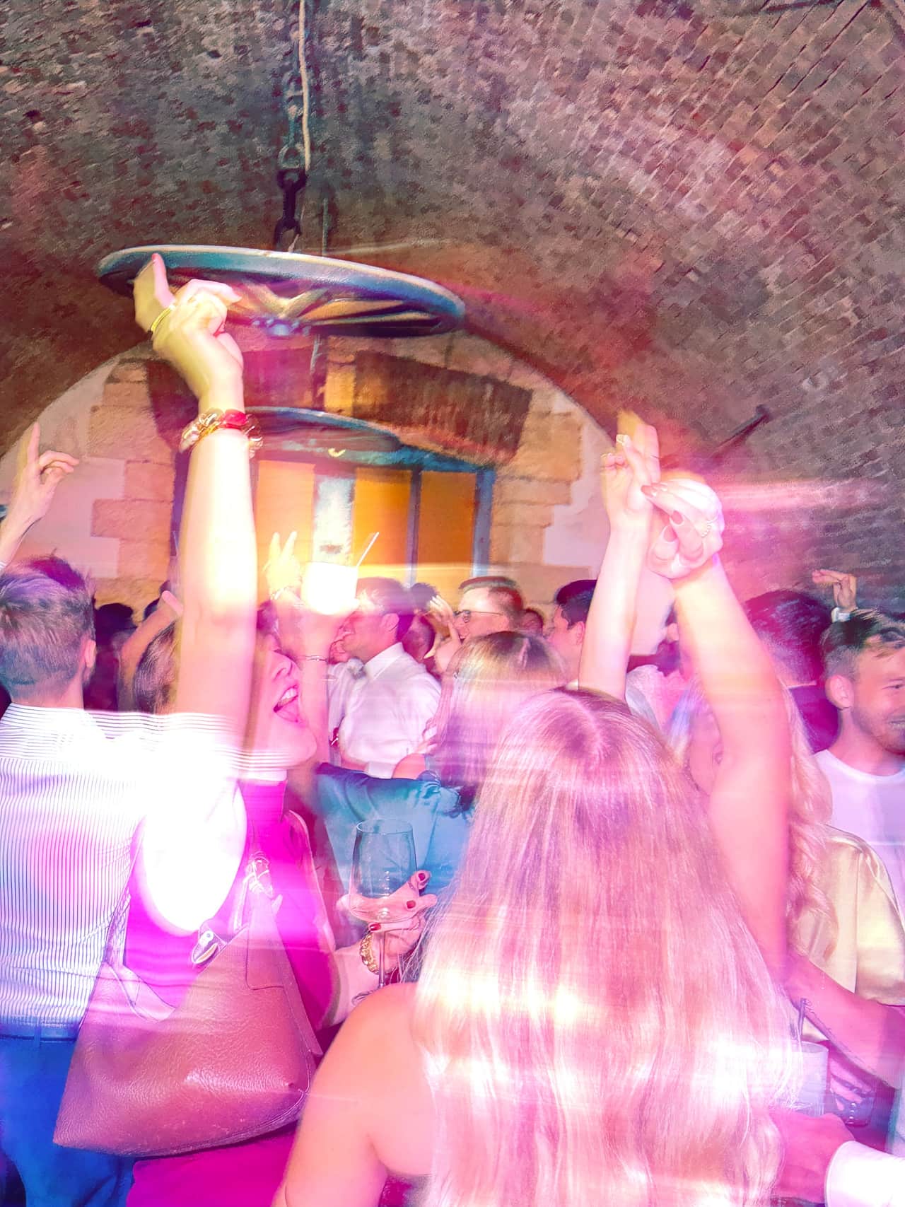 Gäste feiern mit erhobenen Armen in einem Gewölbekeller, das Partylicht taucht alles in Pink
