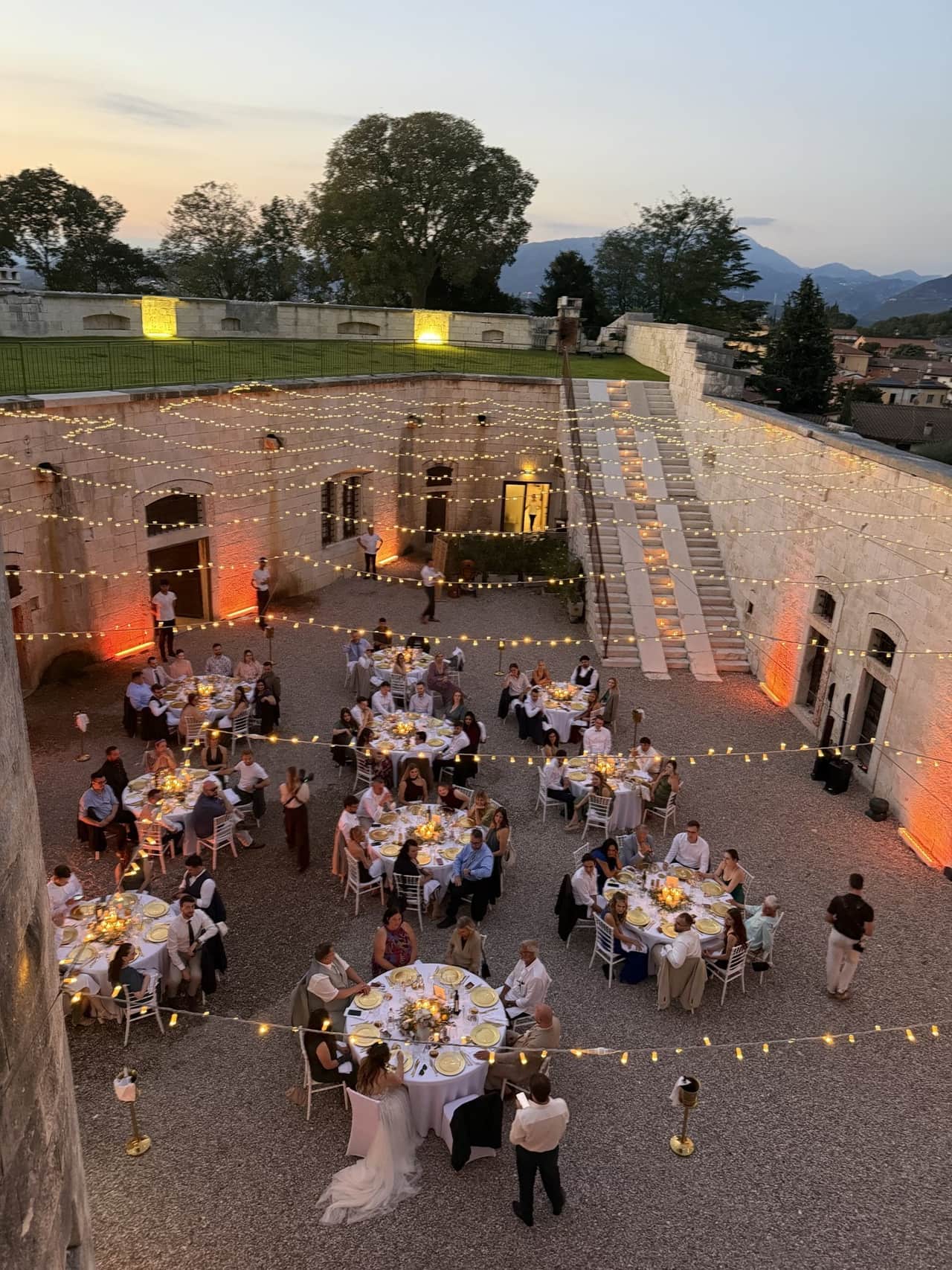 Innenhof von Forte Benedek am Gardasee am Abend, gedeckte runde Tische unter Lichterketten, dahinter Berge